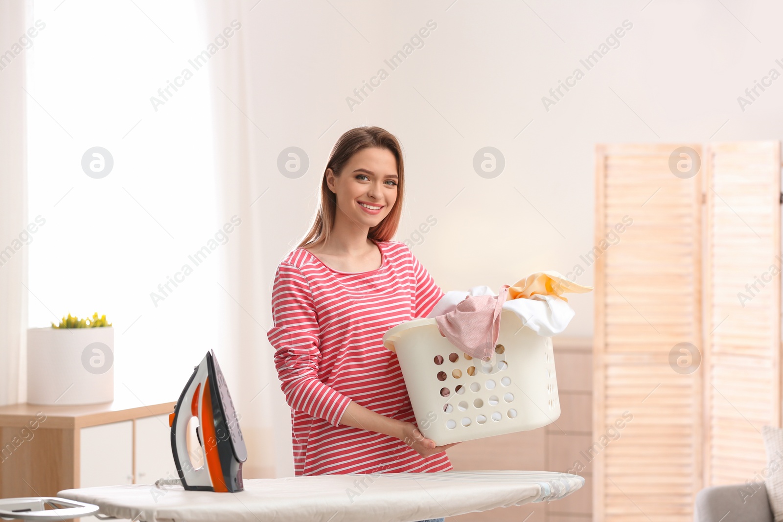 Photo of Young pretty woman holding basket of clean laundry at ironing board indoors