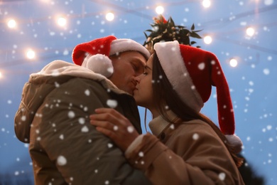 Photo of Happy couple kissing under mistletoe bunch outdoors in snowy evening, low angle view