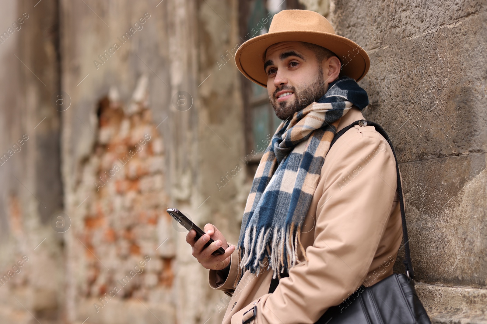 Photo of Smiling man in warm scarf with smartphone near wall outdoors. Space for text