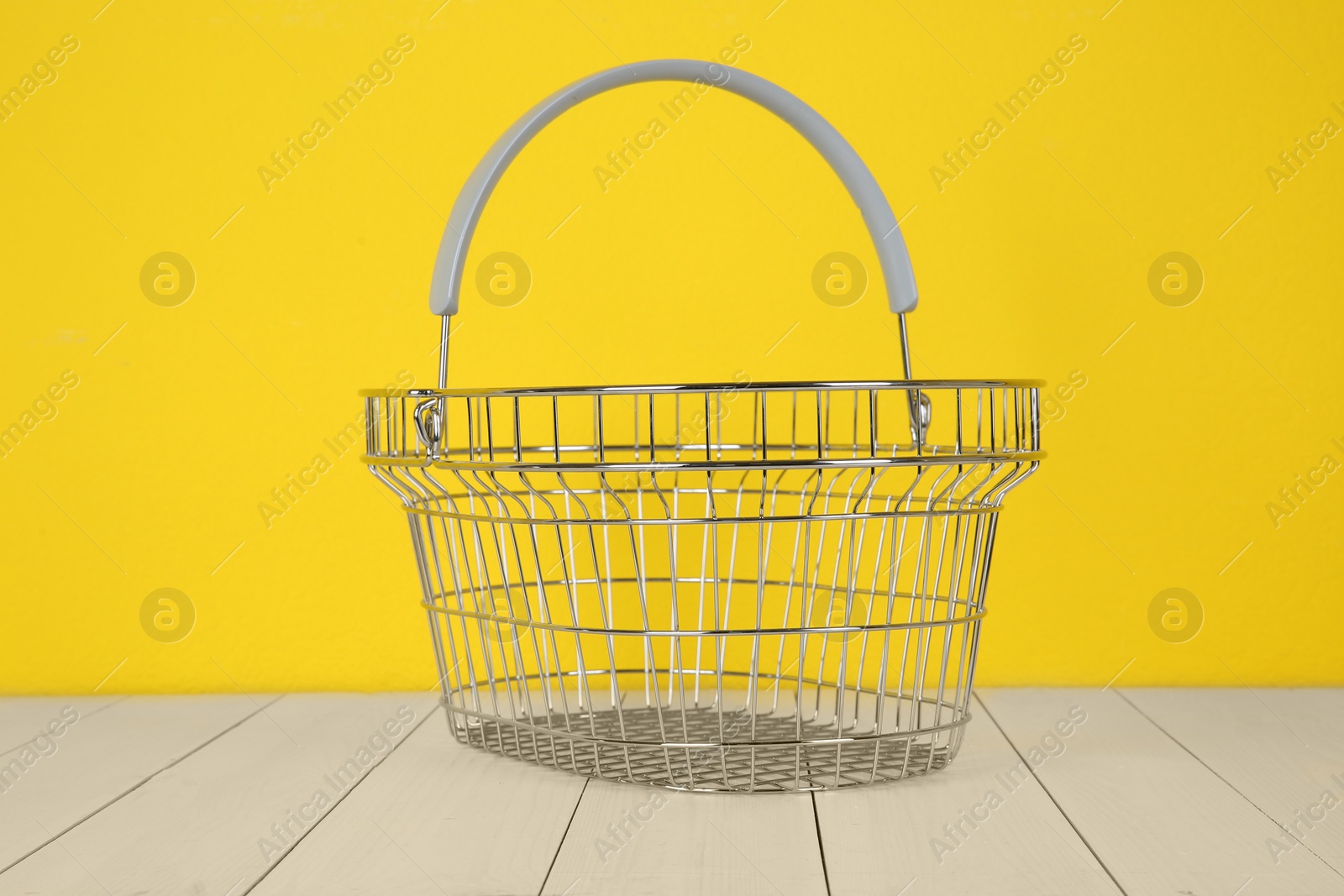 Photo of Empty metal shopping basket on white wooden table against yellow background