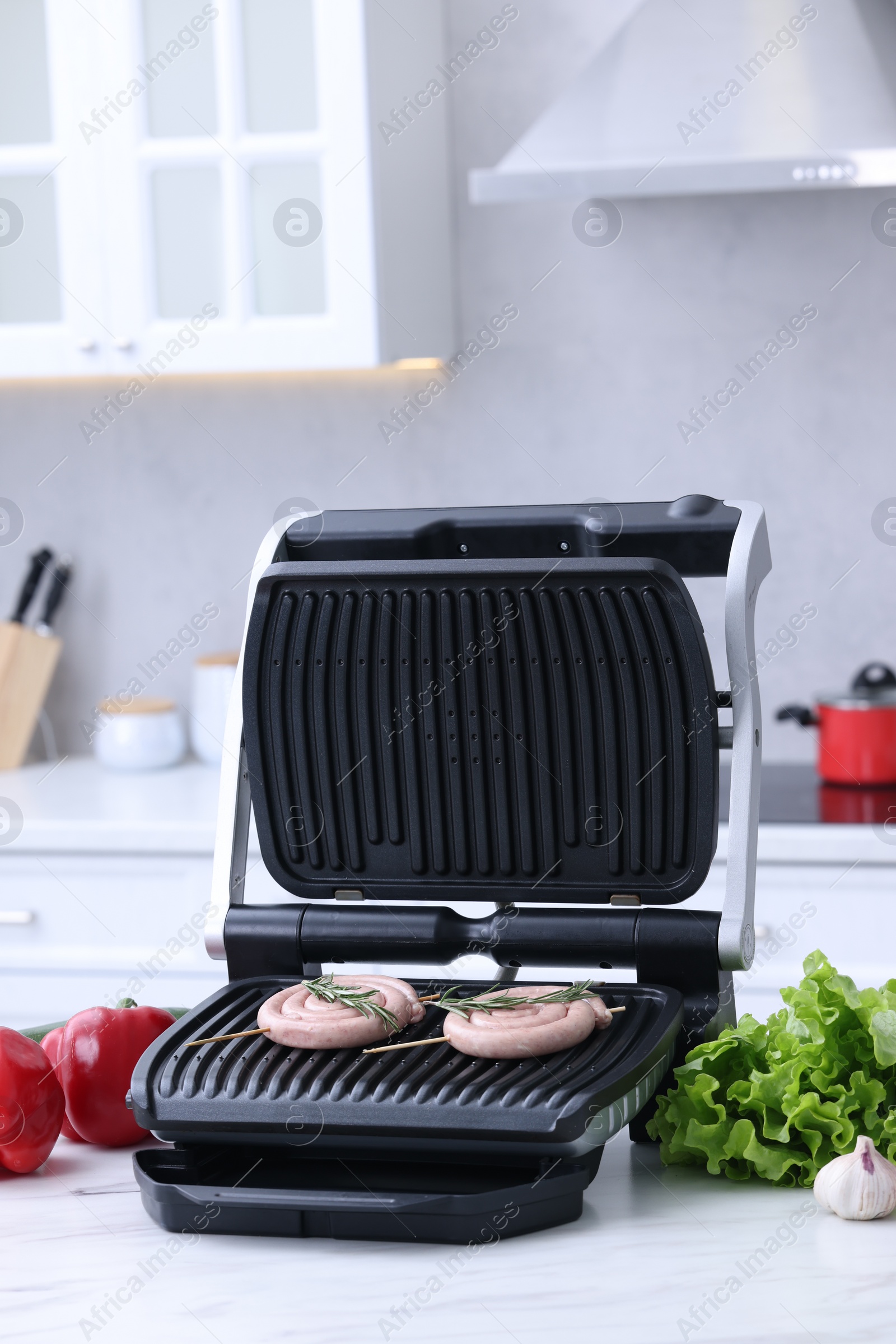 Photo of Electric grill with homemade sausages, rosemary and vegetables on white table in kitchen