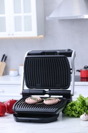 Photo of Electric grill with homemade sausages, rosemary and vegetables on white table in kitchen