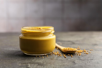 Tasty mustard sauce in jar and spoon with dry seeds on grey textured table, closeup. Space for text