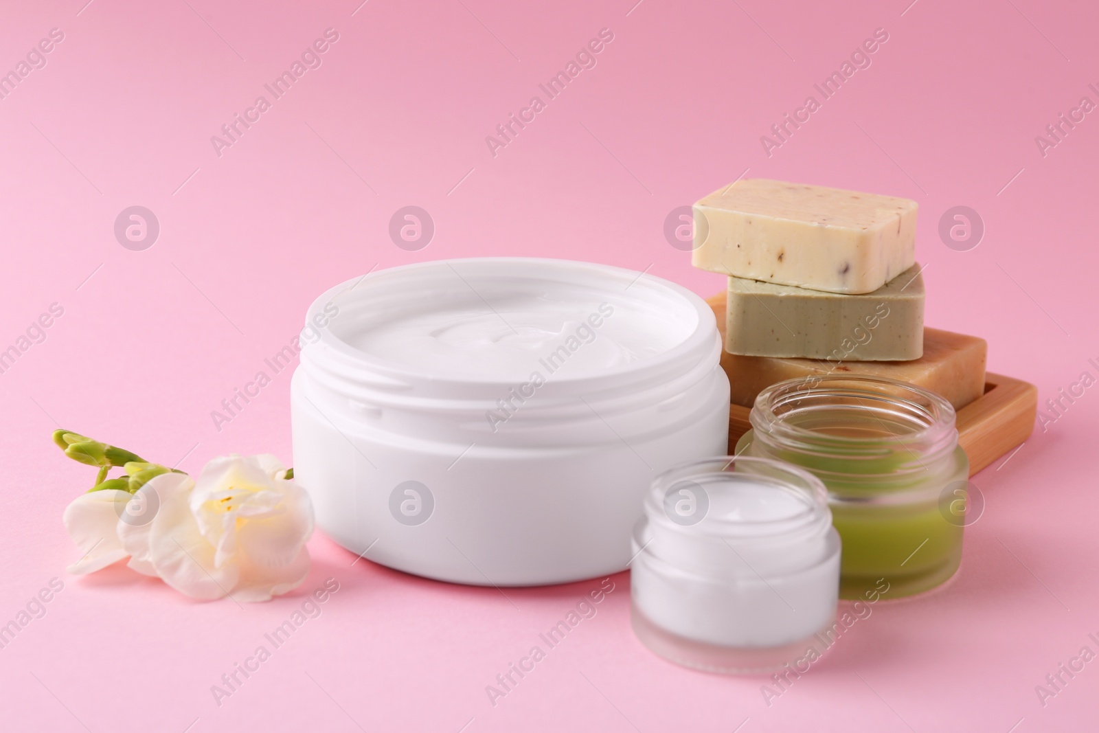 Photo of Moisturizing cream in open jars, soap bars and freesia flower on pink background. Body care products