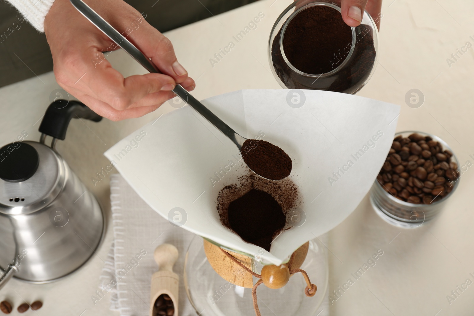 Photo of Making drip coffee. Woman adding ground coffee into chemex coffeemaker with paper filter at white table, closeup