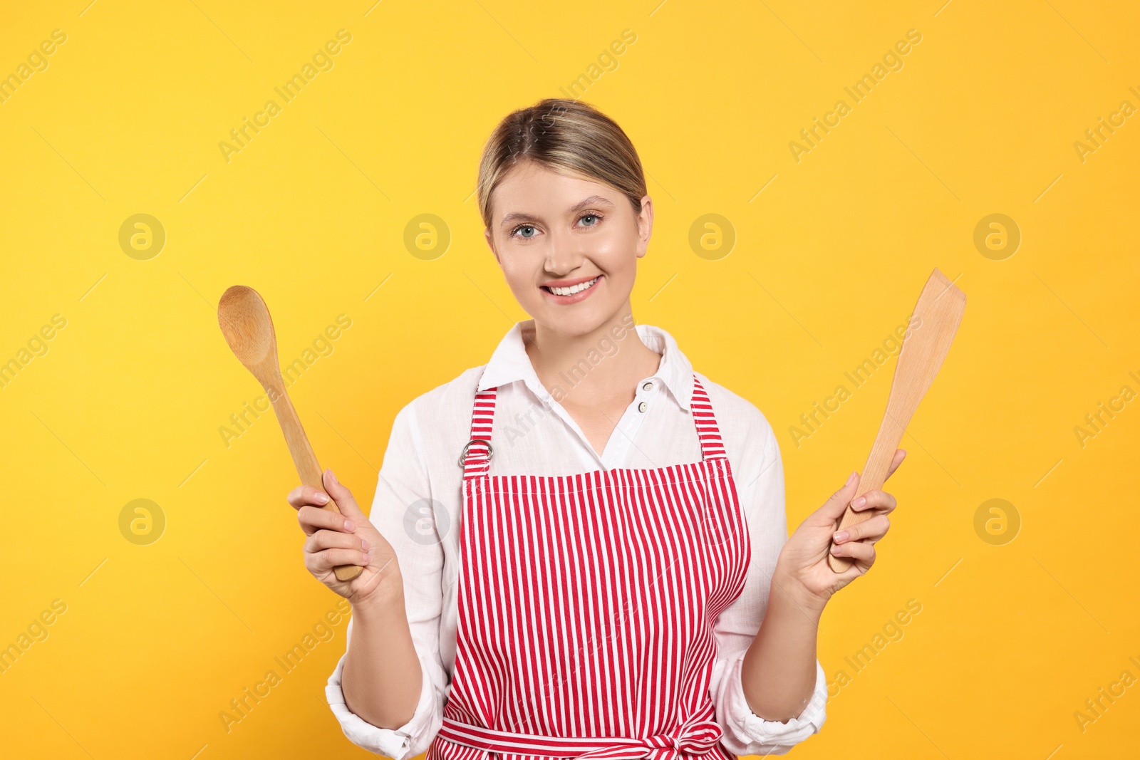 Photo of Beautiful young woman in clean striped apron with kitchen tools on orange background