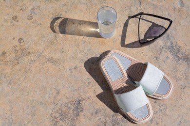 Photo of Stylish sunglasses, slippers and glass of water on stone floor outdoors, space for text. Beach accessories