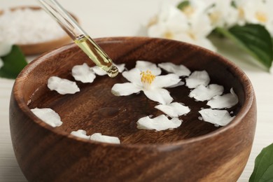 Photo of Dripping jasmine essential oil into bowl with petals on table, closeup