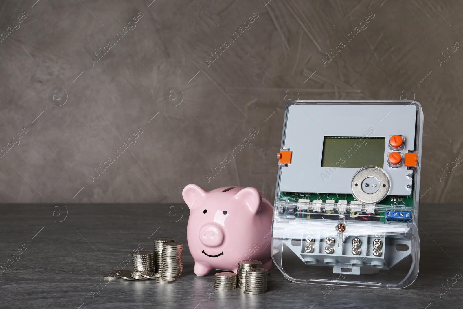 Photo of Electricity meter, piggy bank and coins on grey table. Space for text