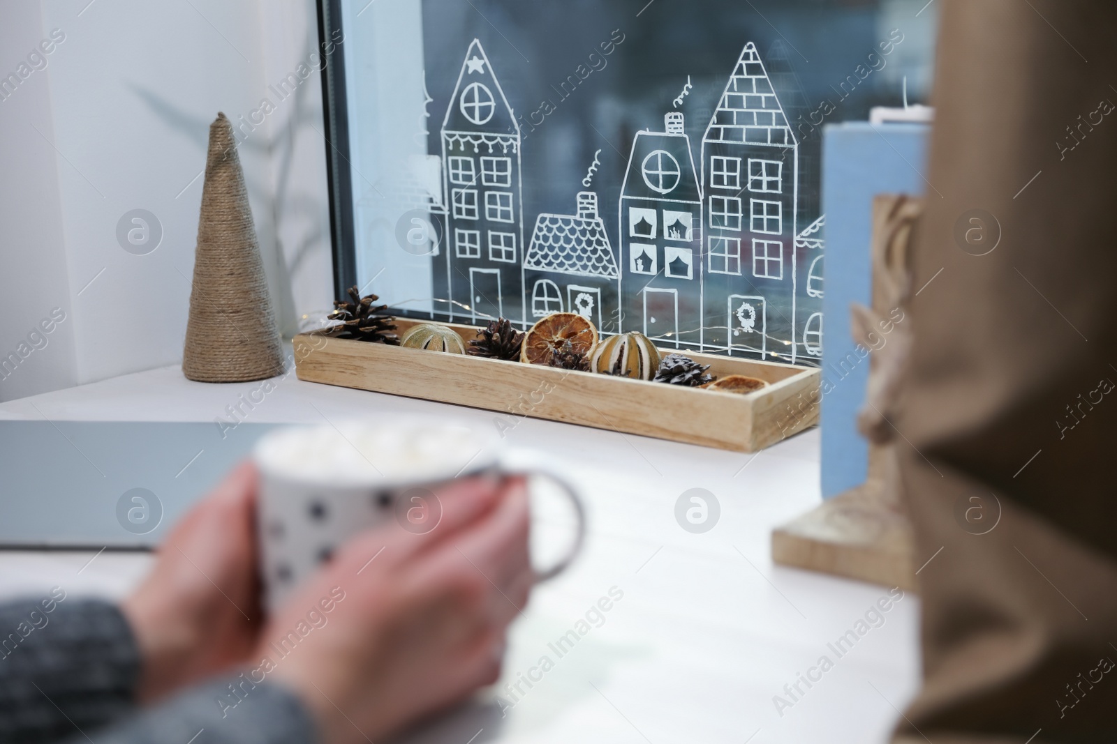 Photo of Woman holding cup of hot drink at white wooden table near window with beautiful drawing indoors, closeup. Christmas decor