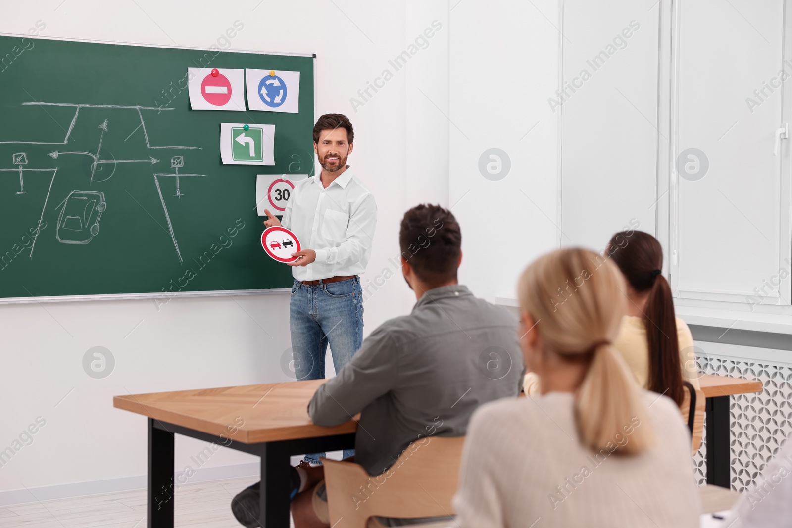 Photo of Teacher showing No Overtaking road sign to audience near chalkboard in driving school