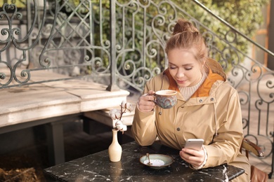 Photo of Young woman enjoying tasty coffee while using mobile phone at table outdoors