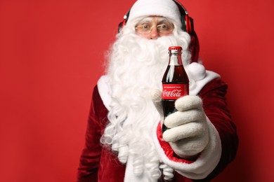 MYKOLAIV, UKRAINE - JANUARY 18, 2021: Santa Claus listening to music with headphones against red background, focus on Coca-Cola bottle in his hand