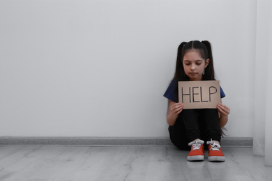 Photo of Sad little girl with sign HELP sitting on floor indoors, space for text. Child in danger