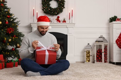 Photo of Happy young man in Santa hat opening Christmas gift at home