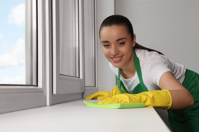 Photo of Happy young woman cleaning window sill with rag indoors