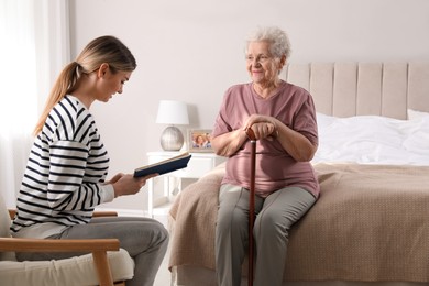 Young caregiver reading book to senior woman in bedroom. Home care service