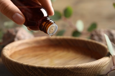 Photo of Woman dripping eucalyptus essential oil from bottle into bowl at wooden table, closeup