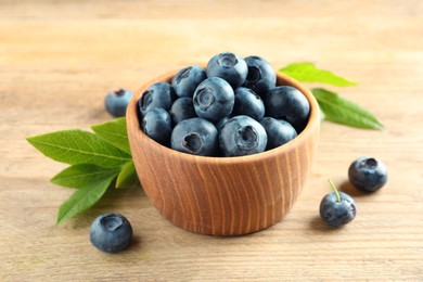 Photo of Bowl of fresh tasty blueberries and leaves on wooden table, closeup