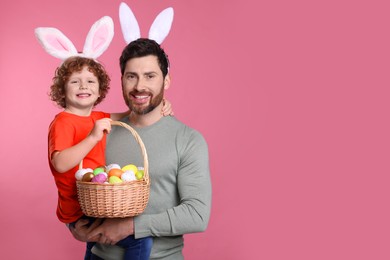 Photo of Happy father and son wearing cute bunny ears headbands on pink background. Boy holding Easter basket with painted eggs, space for text