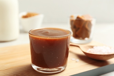 Photo of Glass with tasty caramel sauce and spoon on wooden board