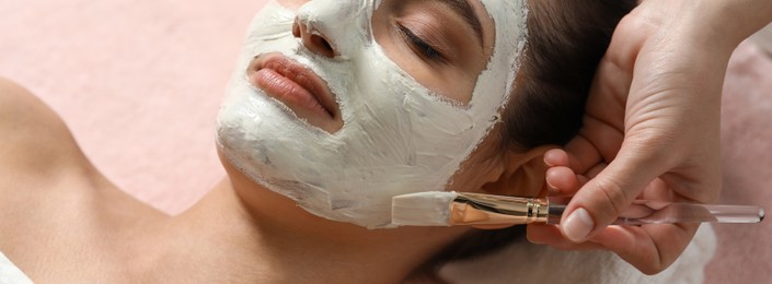 Image of Cosmetologist applying white mask onto woman's face in spa salon, closeup. Banner design