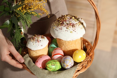 Woman holding basket with traditional Easter cakes, dyed eggs and flowers indoors, closeup