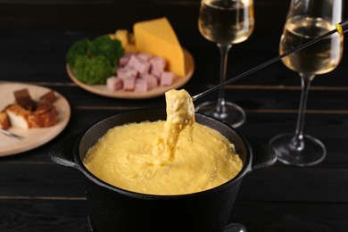 Photo of Dipping piece of ham into fondue pot with melted cheese on black wooden table, closeup