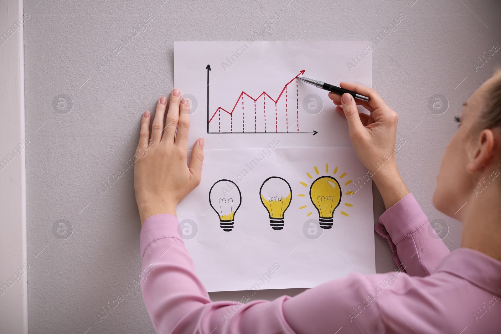 Photo of Woman holding sheets of paper with drawn graphic and lamps near grey wall, closeup. Startup concept