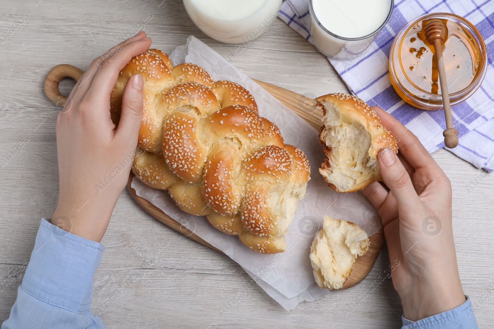 Photo of Woman breaking freshly baked braided bread with sesame seeds at wooden table, top view. Traditional Shabbat challah