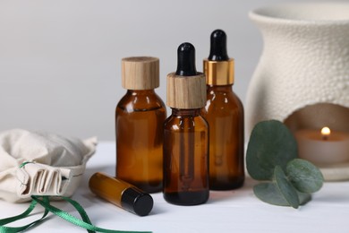 Different aromatherapy products, burning candle and eucalyptus leaves on white wooden table against light background, closeup