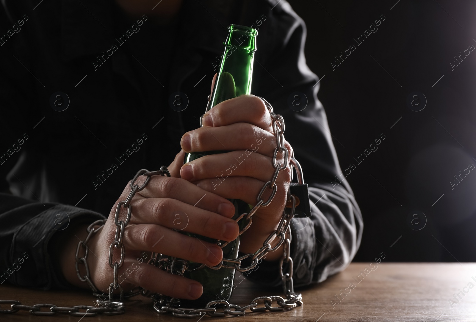 Photo of Alcohol addiction. Man chained with bottle of beer at wooden table, closeup
