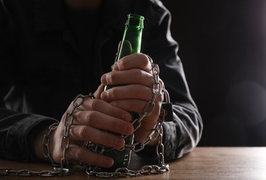 Photo of Alcohol addiction. Man chained with bottle of beer at wooden table, closeup