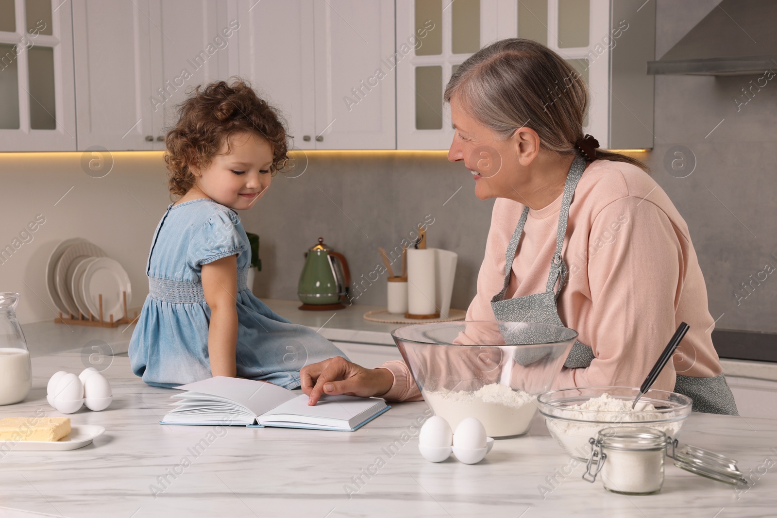 Photo of Cute little girl with her granny cooking by recipe book in kitchen