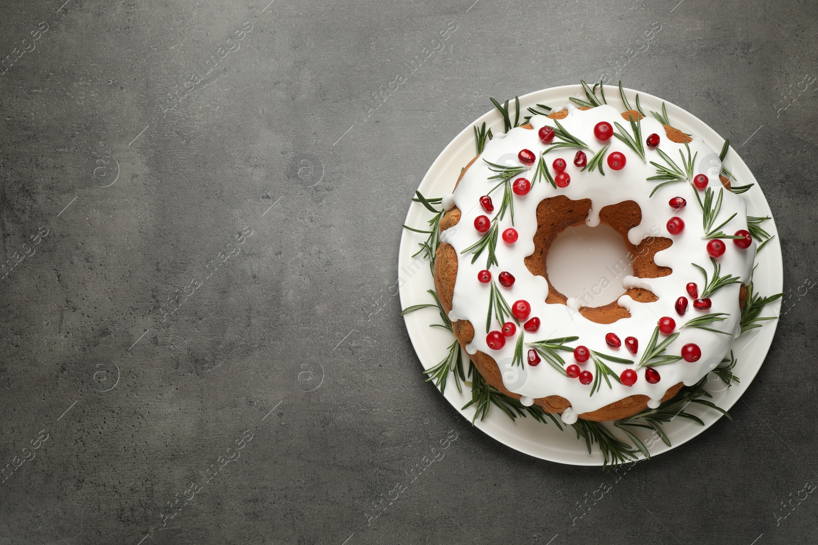 Photo of Traditional Christmas cake decorated with glaze, pomegranate seeds, cranberries and rosemary on grey textured table, top view. Space for text