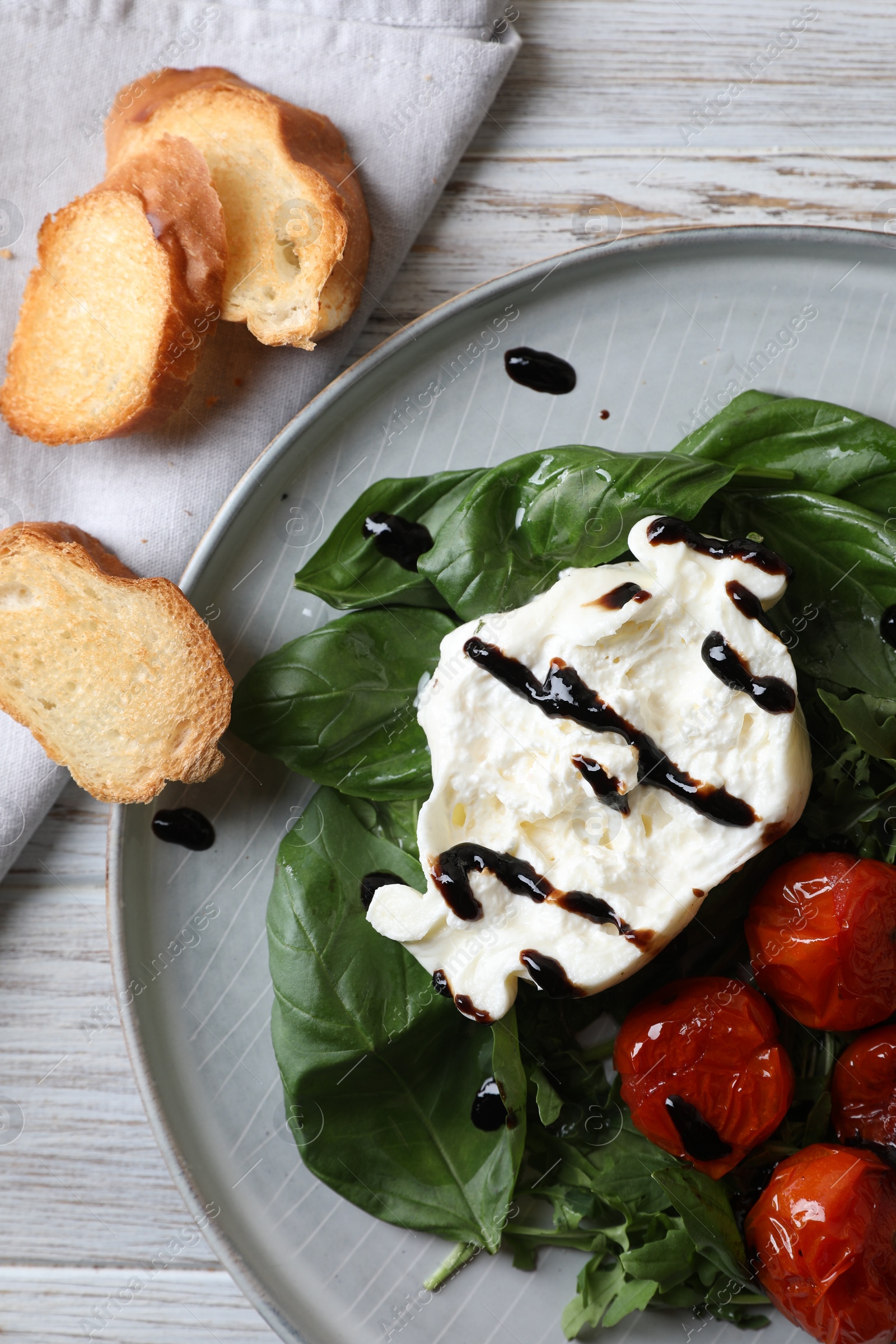 Photo of Delicious burrata cheese served with tomatoes, croutons, arugula and basil on white wooden table, flat lay