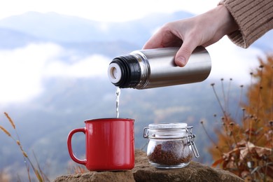 Photo of Woman pouring hot water from thermos into mug with instant coffee in mountains, closeup