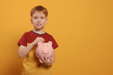 Photo of Cute little boy with ceramic piggy bank on orange background, space for text
