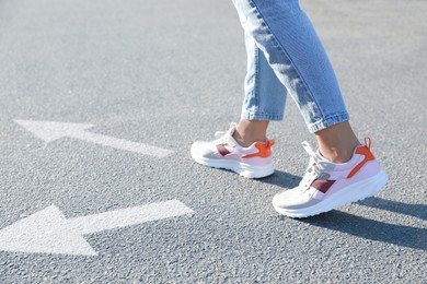 Image of Choice of way. Woman walking to drawn mark on road, closeup. White arrows pointing in different directions