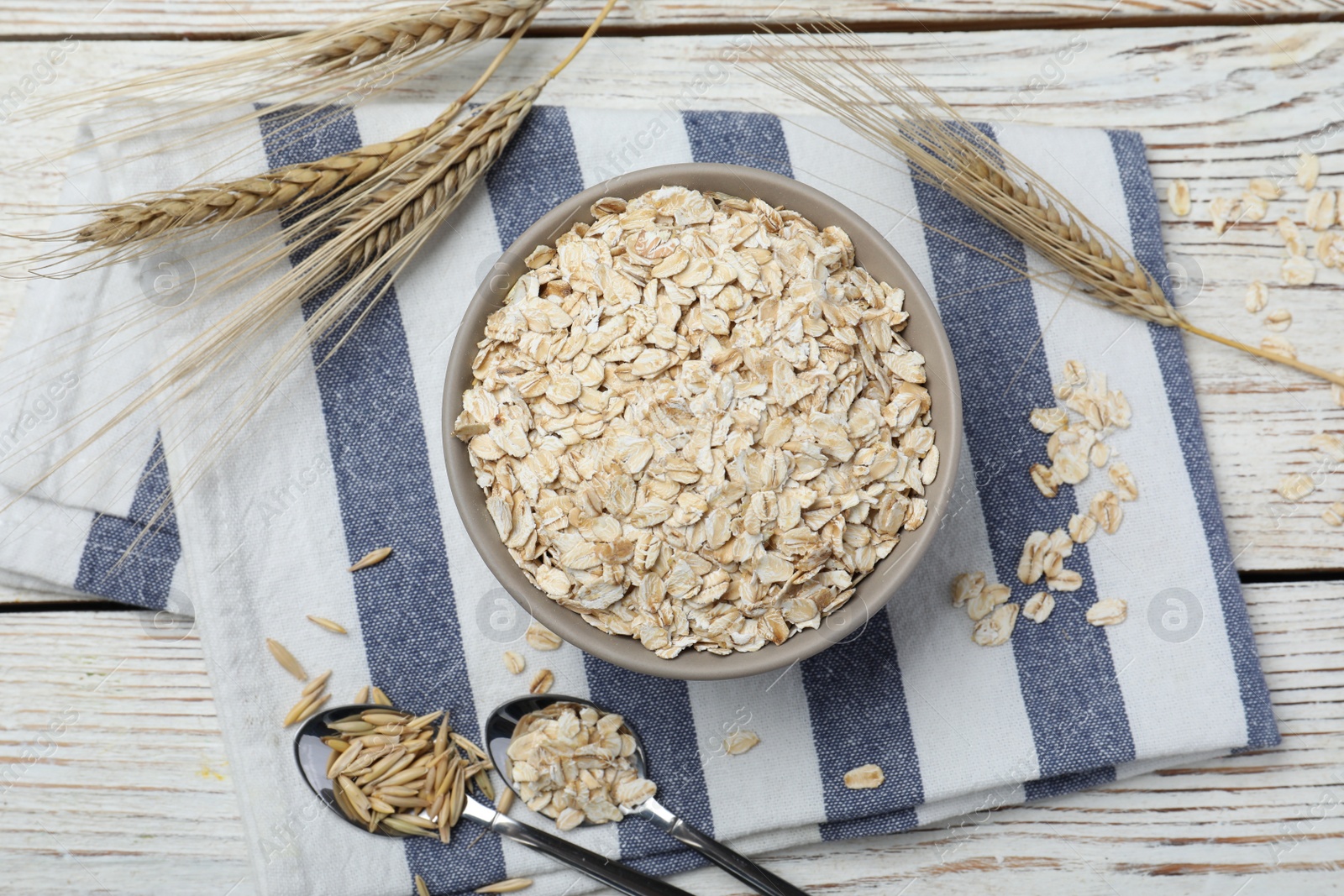 Photo of Bowl of oatmeal, spoons, napkin and spikelets on white wooden table, flat lay