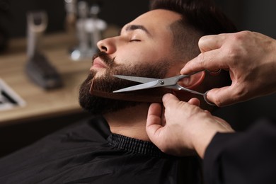 Photo of Professional barber trimming client's beard with scissors in barbershop, closeup