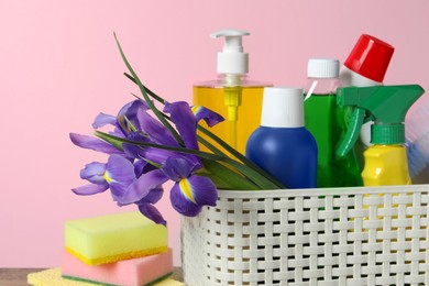 Photo of Spring cleaning. Basket with detergents, flowers and sponges on table against pink background, closeup
