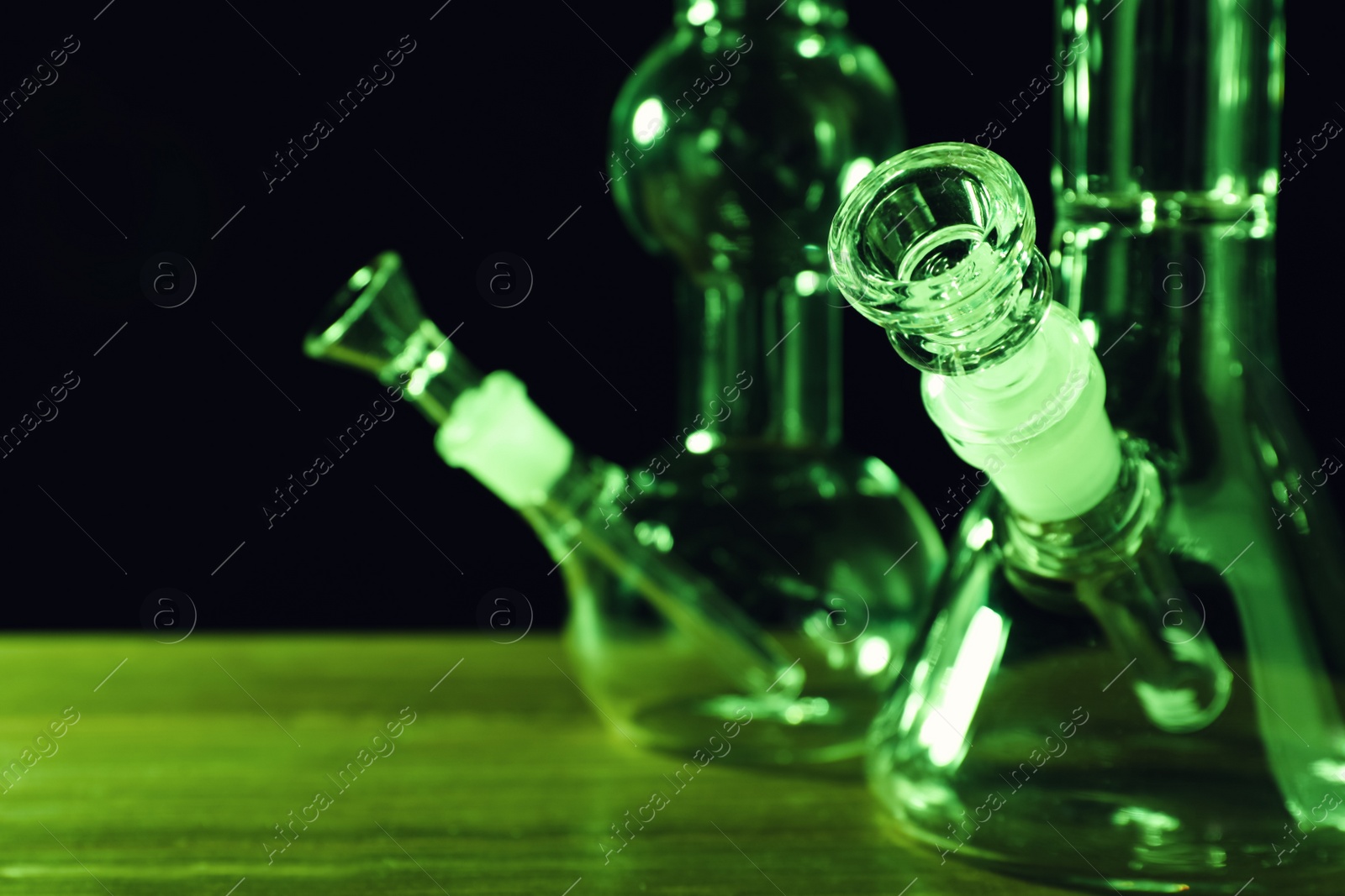 Photo of Glass bongs on wooden table against black background, toned in green. Smoking device