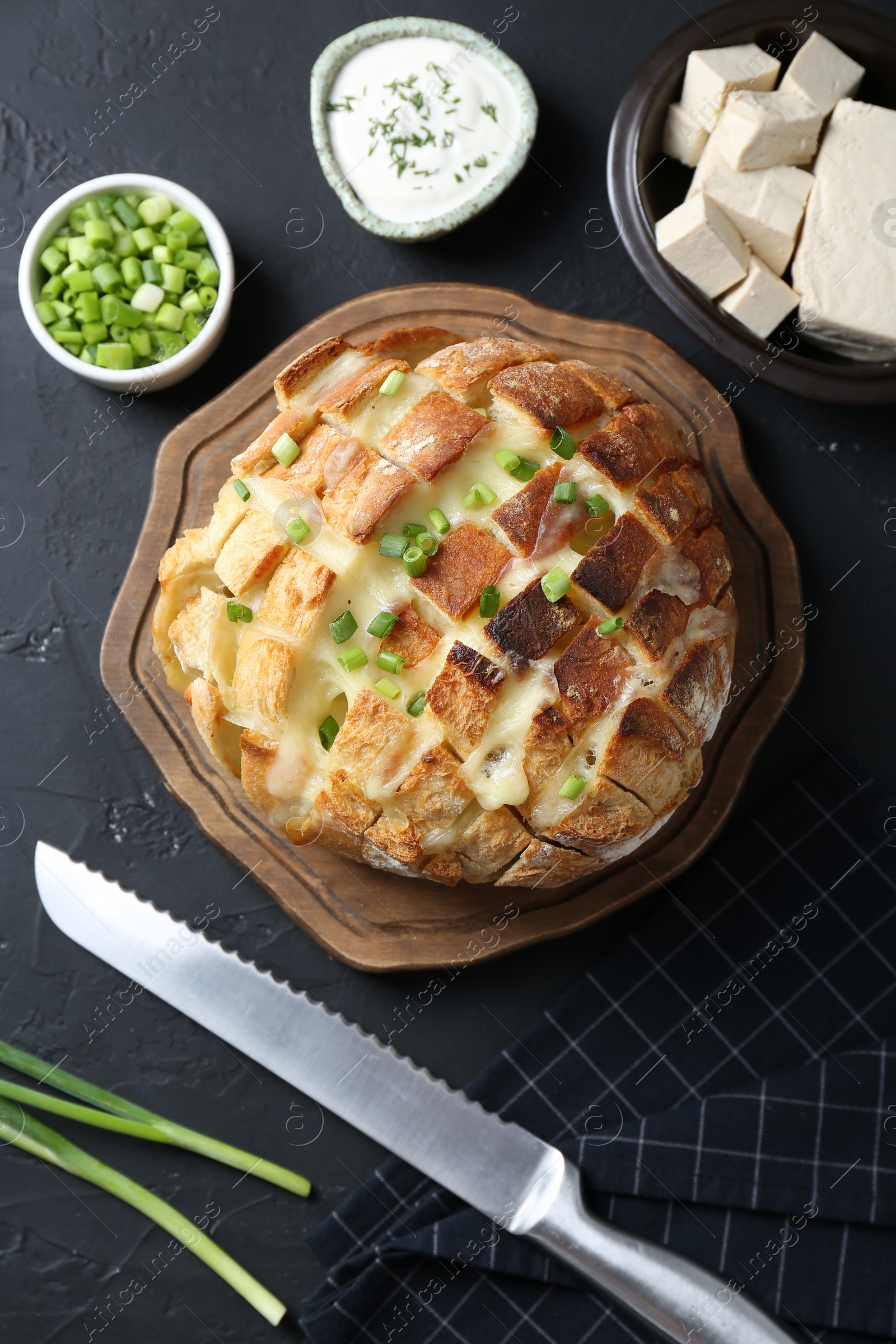 Photo of Freshly baked bread with tofu cheese, green onions, sauce and knife on black table, flat lay