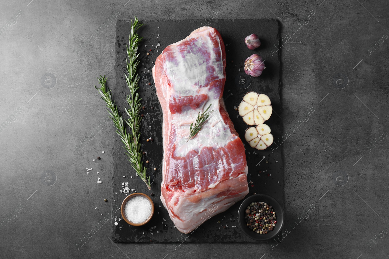 Photo of Piece of raw pork belly, rosemary, garlic and spices on grey textured table, top view