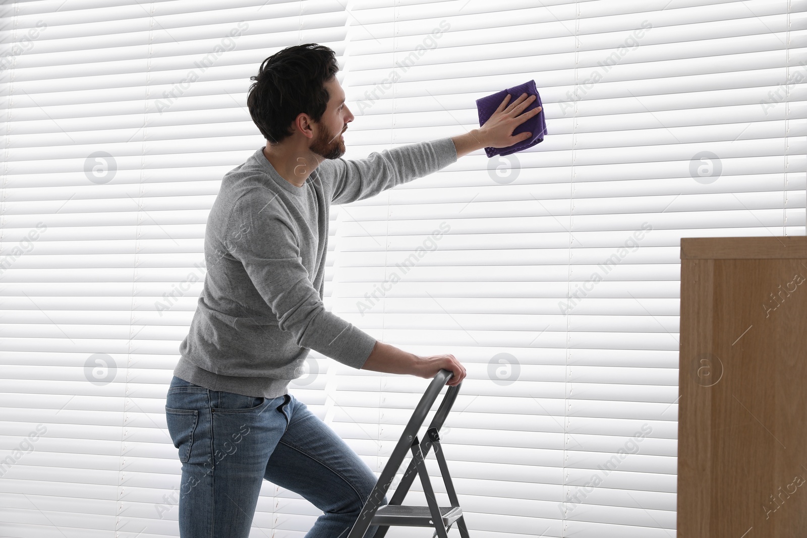 Photo of Man on metal ladder wiping blinds indoors