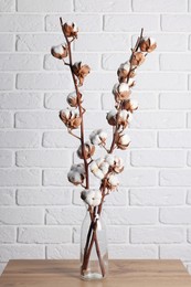 Cotton branches with fluffy flowers in vase on wooden table near white brick wall