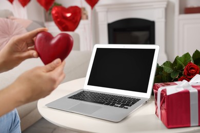 Photo of Valentine's day celebration in long distance relationship. Woman holding red wooden heart while having video chat with her boyfriend via laptop indoors, closeup