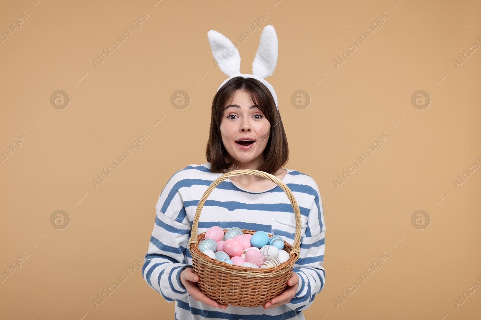 Photo of Easter celebration. Surprised woman with bunny ears and wicker basket full of painted eggs on beige background