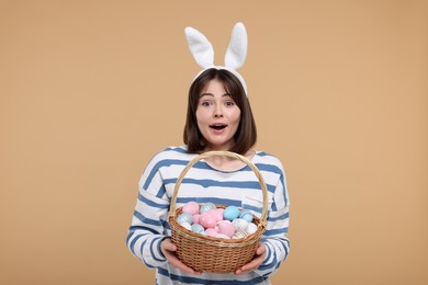 Easter celebration. Surprised woman with bunny ears and wicker basket full of painted eggs on beige background